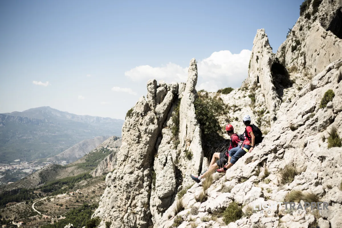 Two climbers sitting side to side on a slab, looking toward a rocky formation resembling three fins