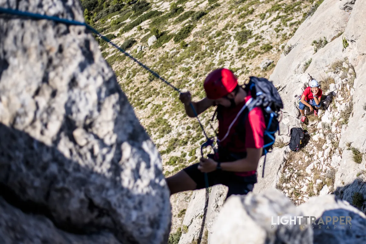 A climber abseiling in foreground, with another, smiling climber watching it underneath