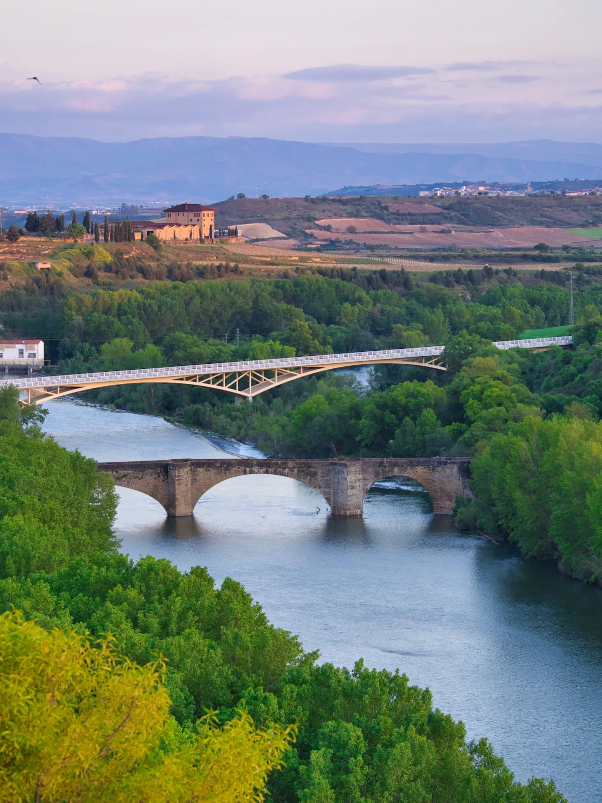 A river, a medieval stone bridge, a modern bridge and a winery building in the background