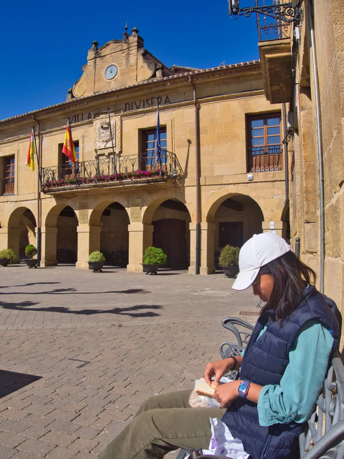 A woman sitting on a bench making a sandwich with town hall in the background