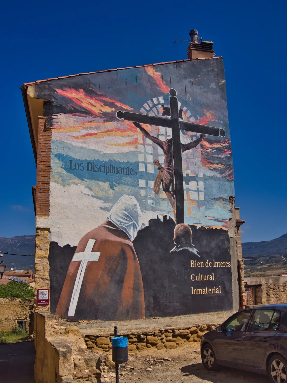 A mural on a wall of a building depicting a man dressed in a brown tunic, Jesus Christ on a cross and sky with fire with ‘Los Disciplinantes’ written on it