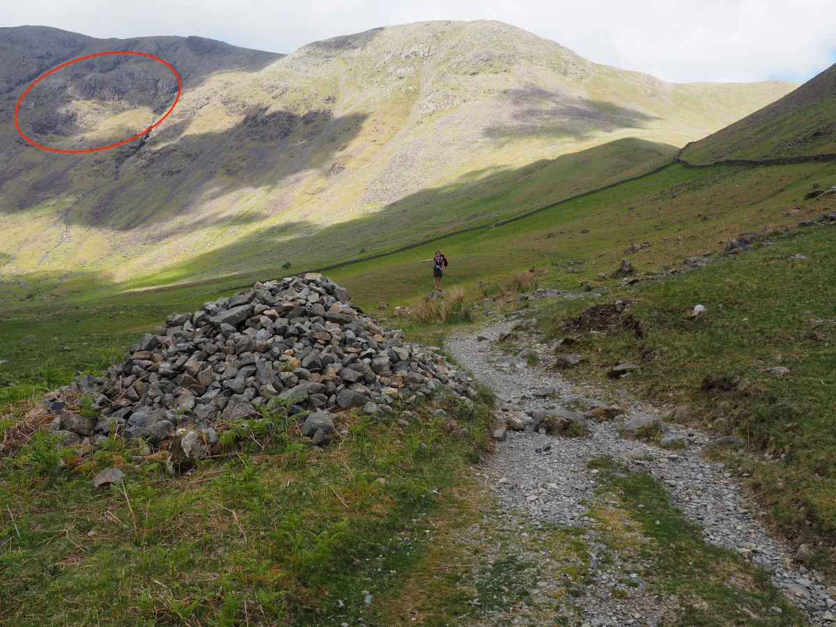 A pile of gravel next to a path; a climber showing the way behind it; Wistow Crags circled in the background