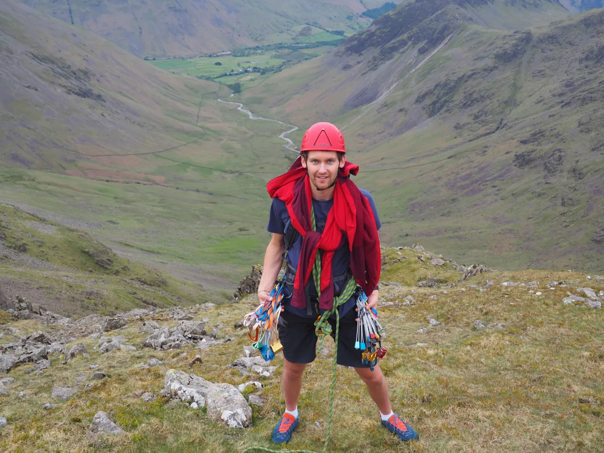 A happy climber in a helmet with rope coiled around his torso, showing his climbing protection: nuts, cams (friends) and hexes